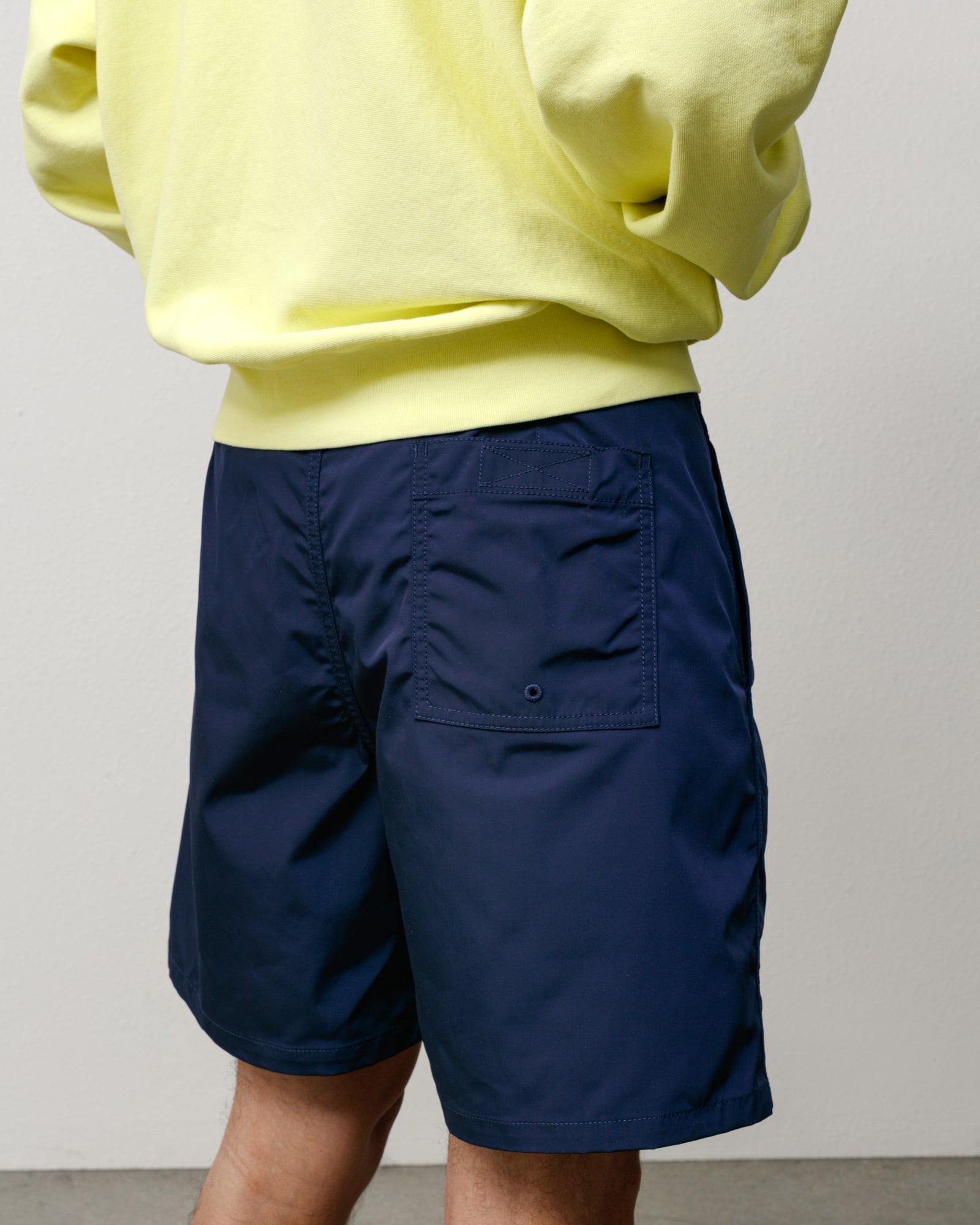 Blue board shorts, rear view showing patch pocket with drainage eyelet and smooth woven fabric.
