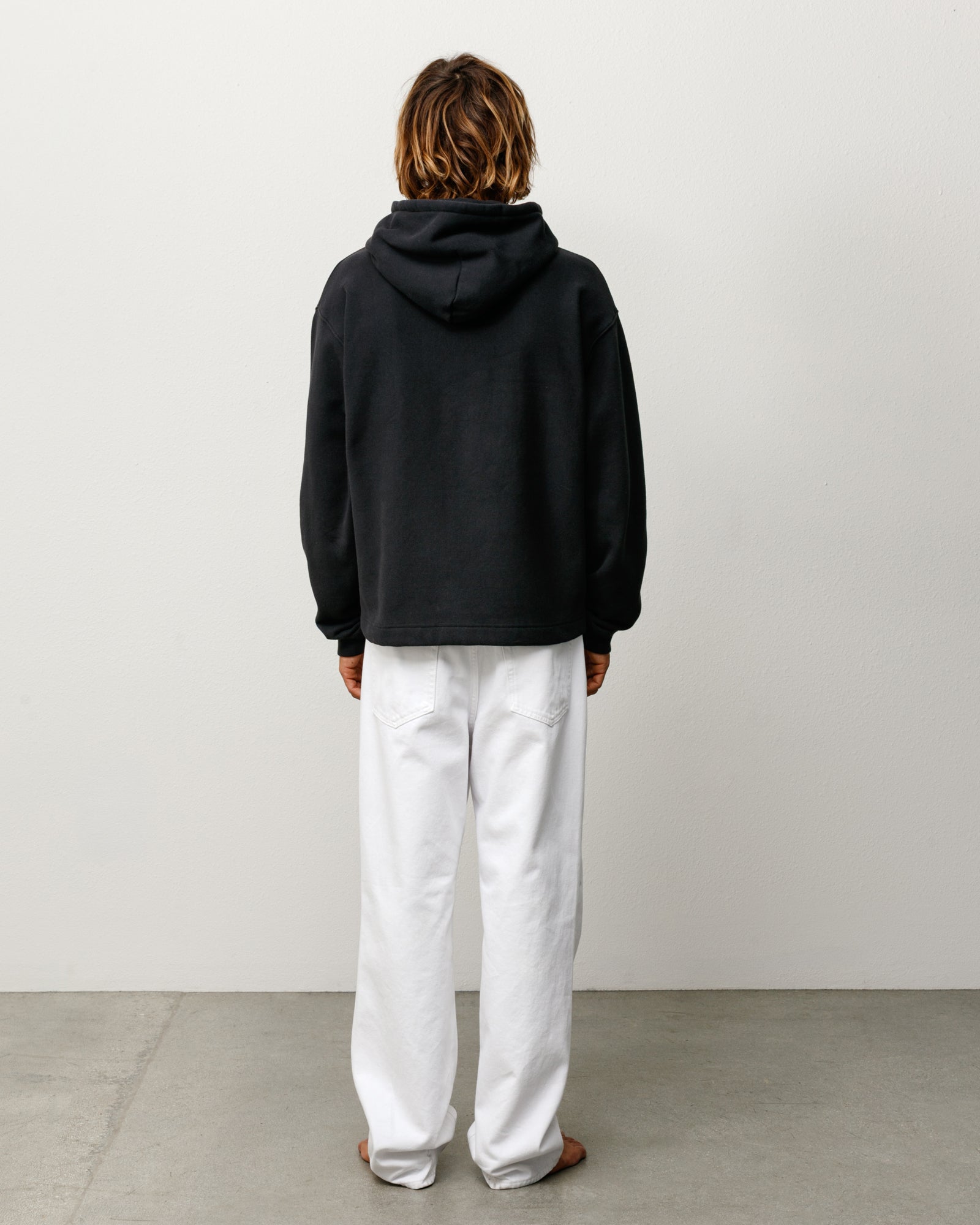 Black pullover hoodie with long sleeves, back view on model wearing white trousers