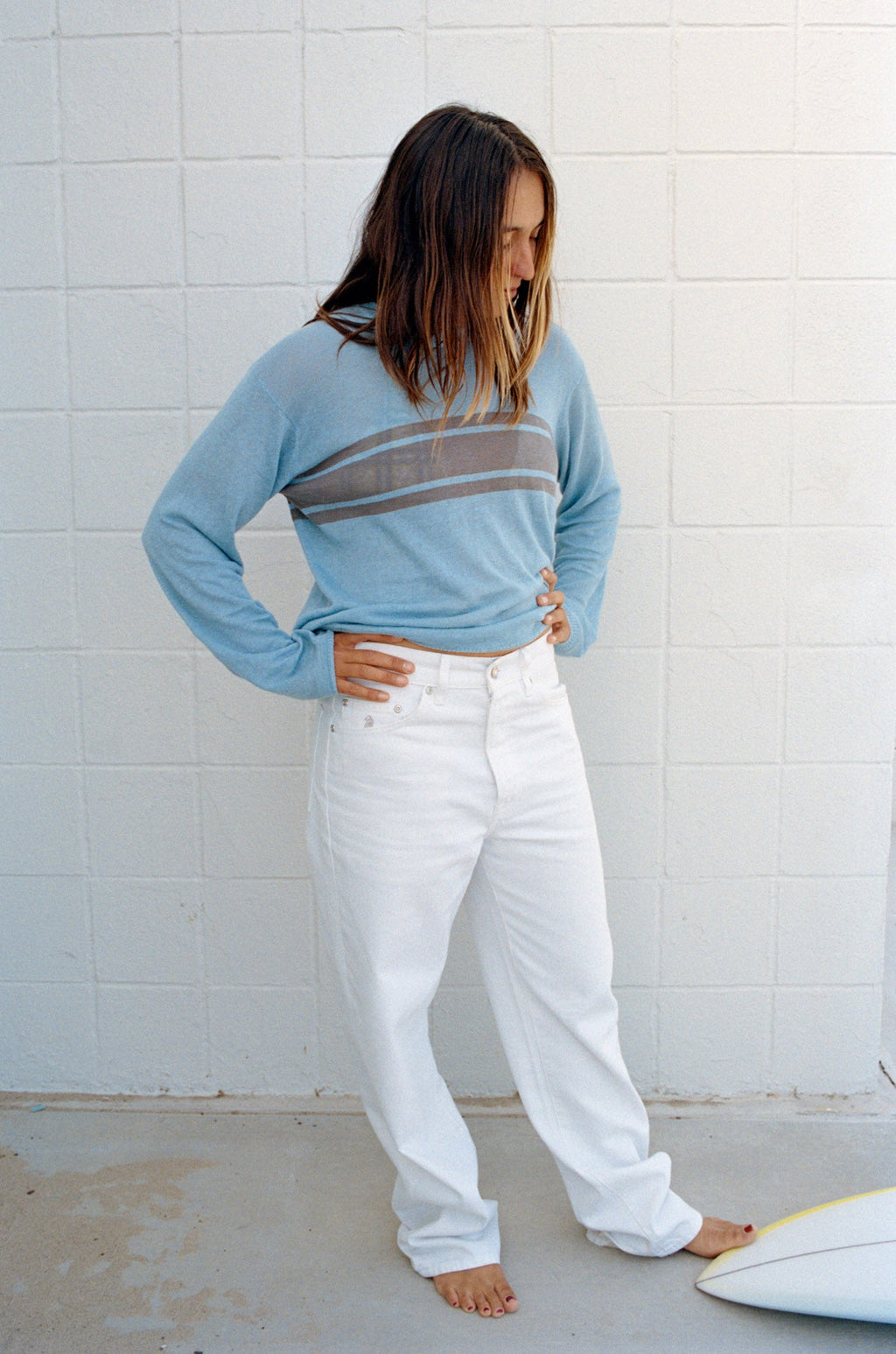 Model in the Lightweight Striped Sweater in Blue and Big Ol' Jean Denim in White standing against a white brick wall.