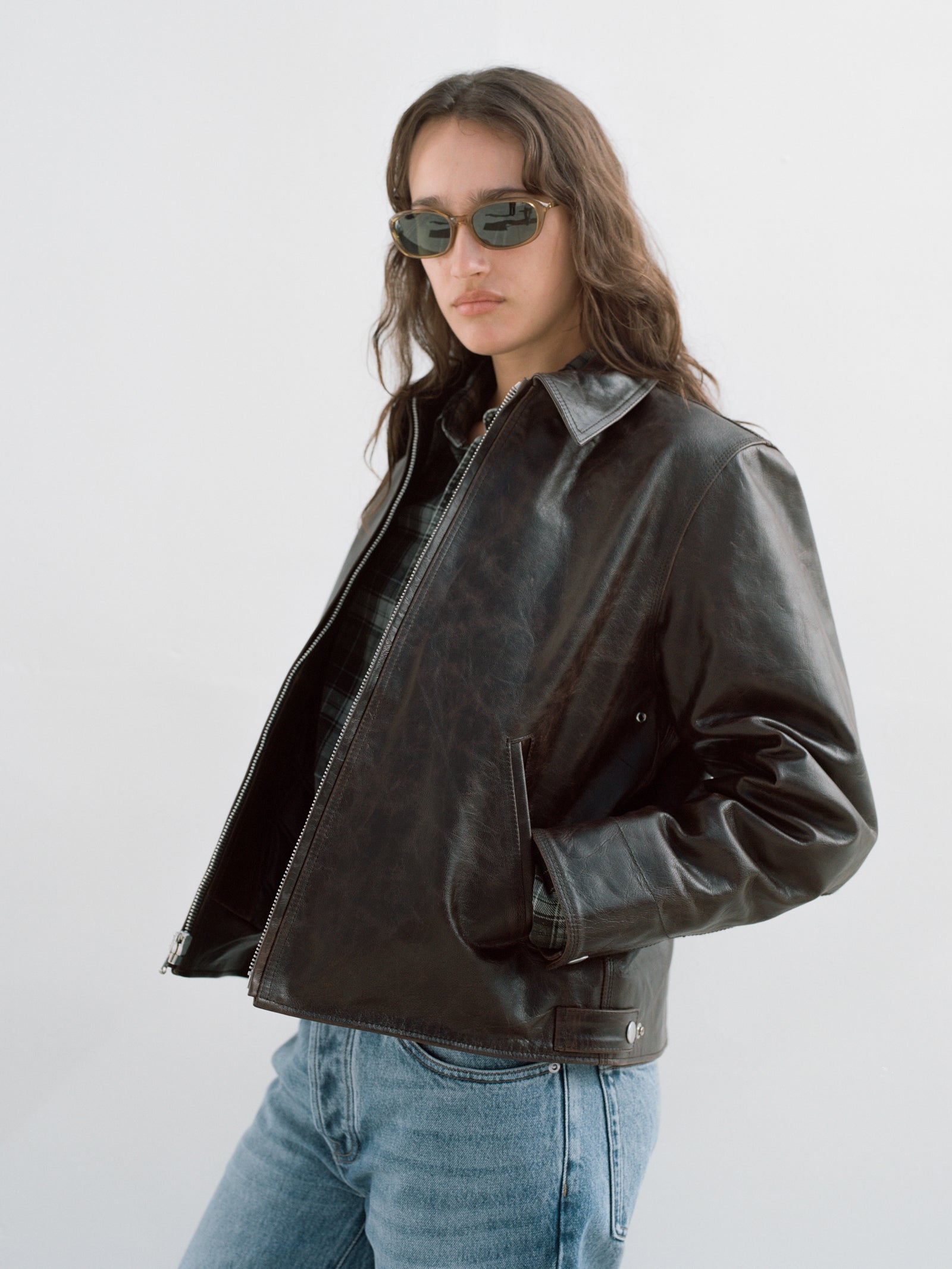 Person wearing the Garage Leather Jacket In Dark Brown, green-tinted sunglasses, and the indigo stone wash Slim Jean Denim against a neutral background.