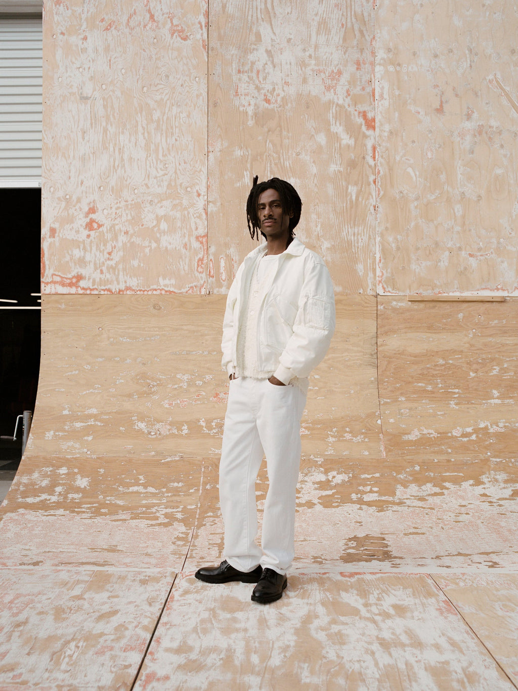Full-body portrait of a person wearing the Mock Neck Flight Bomber in White layered over the Textured Cardigan in Off White and the Slim Jean Denim in White with black shoes. They stand with one hand in their pocket in front of a textured plywood wall and floor backdrop, facing the camera with a calm expression.