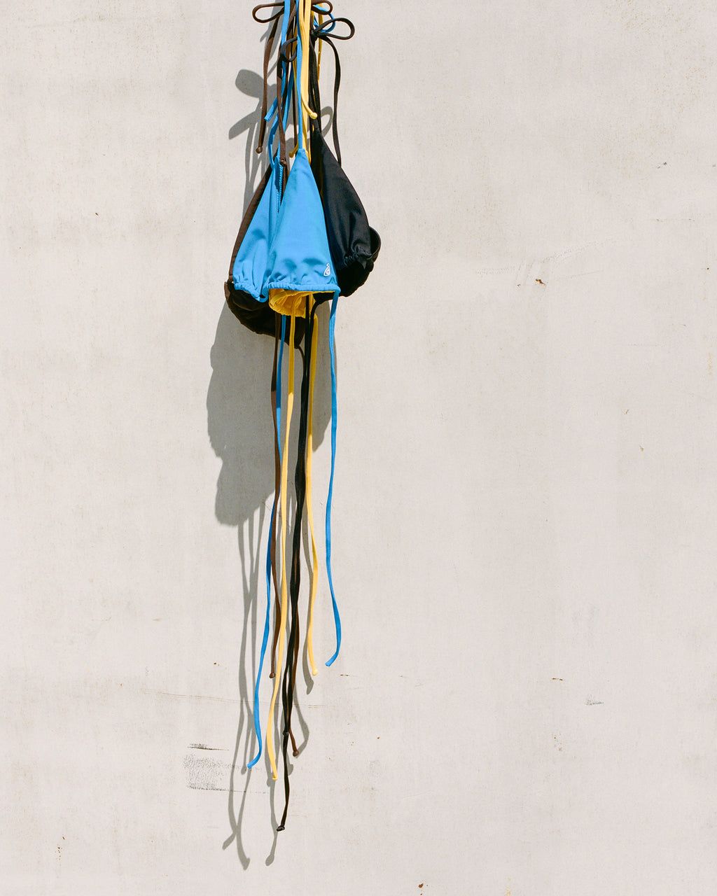 Triangle Bikini Tops Turquoise, Brown, Orange, and Black hanging from their straps against a plain, light-colored wall. 