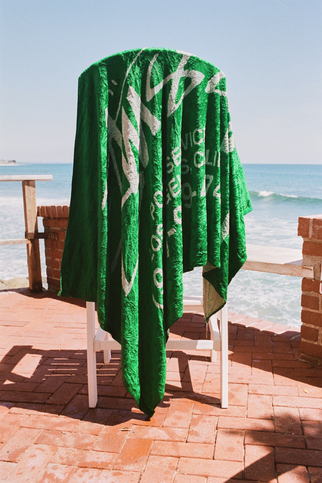 The Pool Service Beach Towel in Green draped over a chair on a brick patio overlooking the ocean.