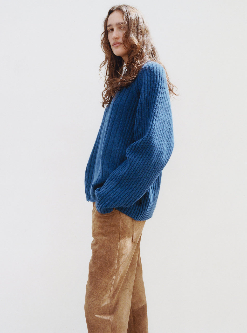 A model with long wavy hair wears the Merino Roll Neck Sweater in Blue and New Classic Pant Suede in Tan against a plain white background.