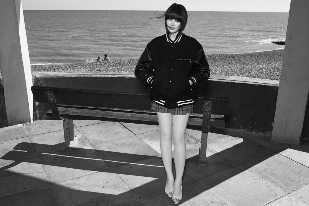 A black-and-white portrait of a woman standing in front of a wooden bench by the ocean, wearing the JUNYA VARSITY JACKET and a plaid skirt.