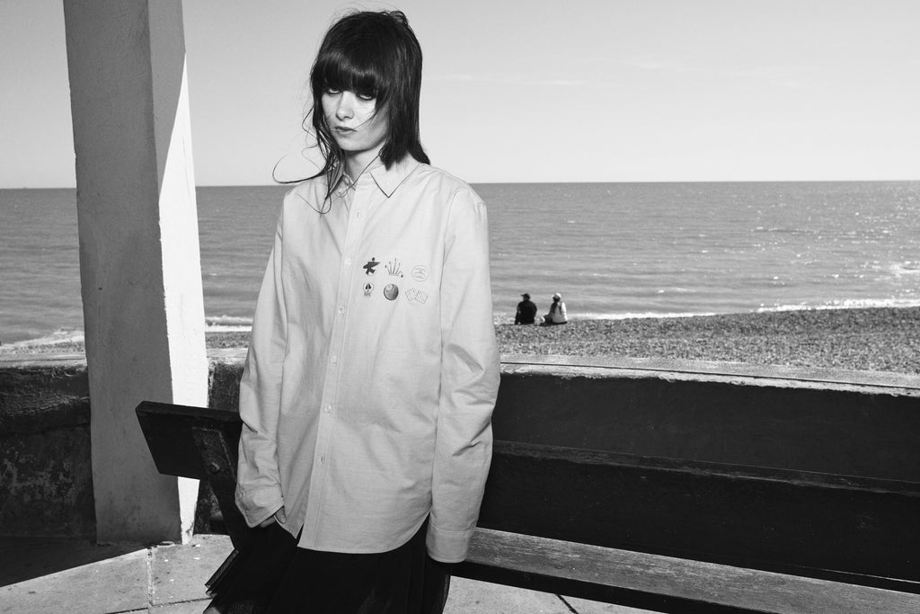 A black-and-white portrait of a woman leaning against a wooden bench outdoors, wearing the JUNYA BUTTON DOWN SHIRT in SAXE BLUE.