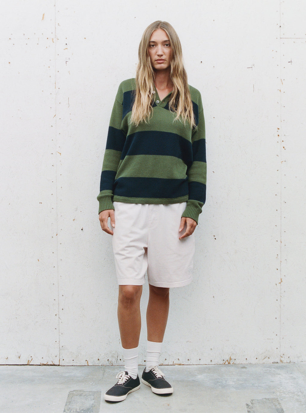 A woman with long blonde hair standing in front of a textured wall, wearing the PIQUE KNIT STRIPE RUGBY SWEATER in OLIVE  and white shorts.