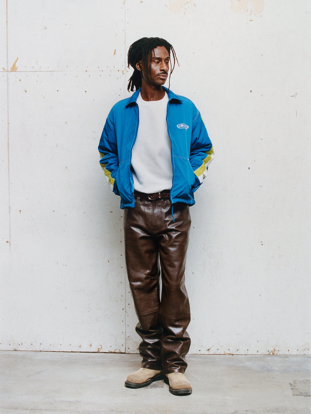 Full-body shot of a model with dreadlocks wearing the Offshore Jacket in Royal over a white t-shirt with the New Classic Pant Leather in Brown against a textured wall.