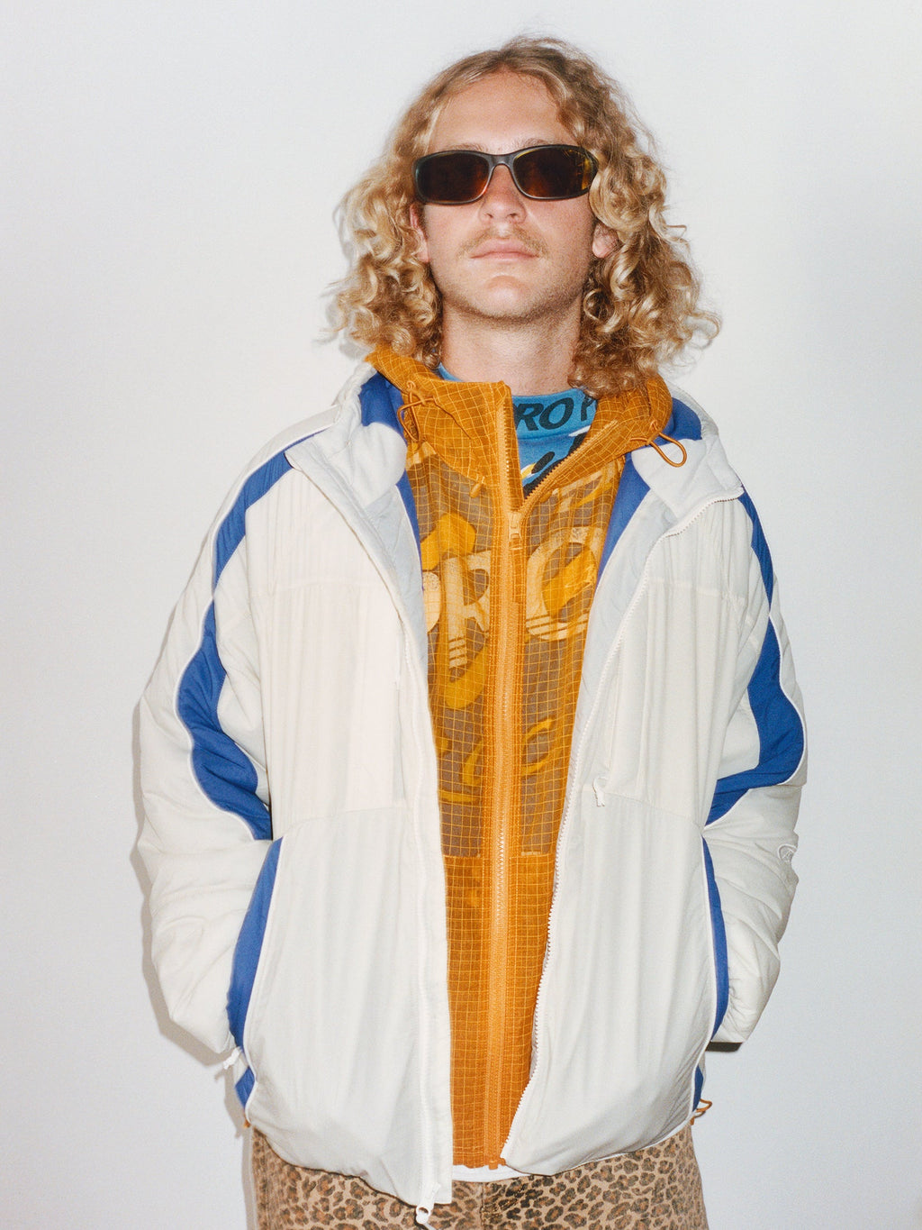 Medium close-up of a model wearing dark sunglasses, the Rally Insulated Jacket in Bone, and leopard-print pants against a plain white background.