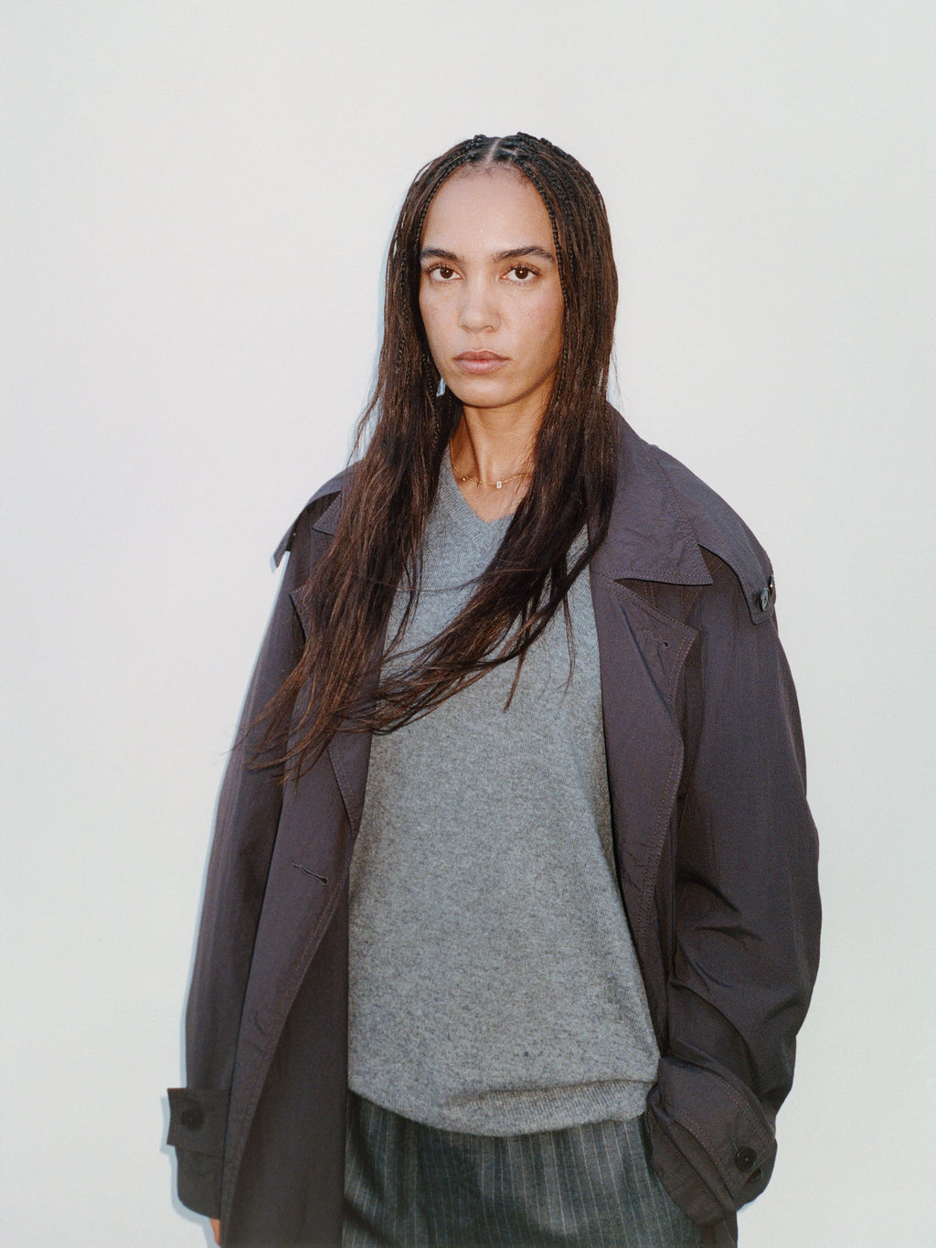 Portrait of a model with long braided dark hair wearing the Ultra Lightweight Nylon Trench in Brown layered over a light grey V-neck sweater and pinstripe trousers against a plain off-white backdrop.